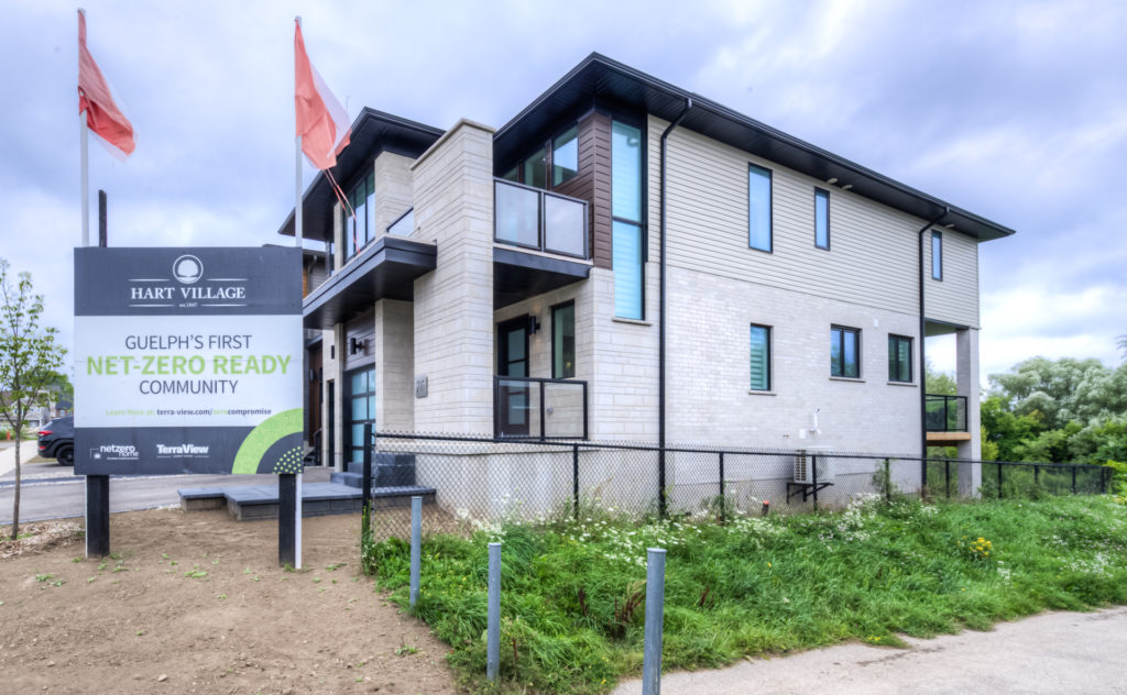 Modern two-story building with balconies, beige and grey siding, large windows, and red flags. A sign in front reads “Guelph’s First Net-Zero Ready Community” by Hart Village, highlighting the neighbourhood’s commitment to net zero homes. Grass and path in the foreground emphasize this innovative example of net zero communities.