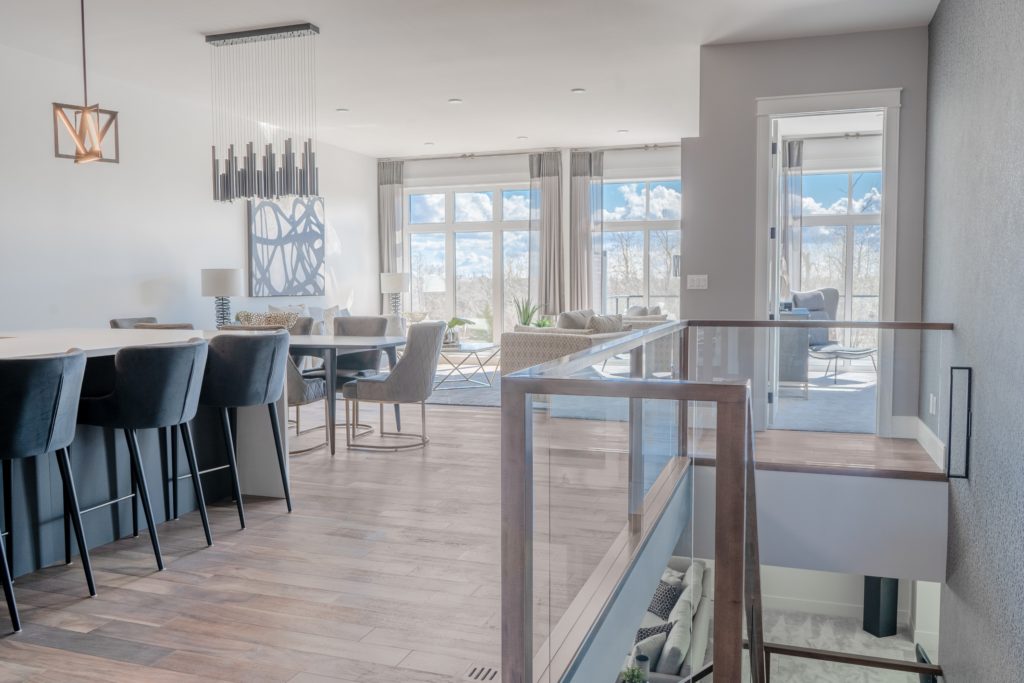 Modern open-concept living space with large windows, light wood floors, a dining area with high-back chairs, and a bright living room with neutral decor. Natural light fills the room through the expansive windows, creating an inviting atmosphere that highlights the sustainable design found in many net zero homes.