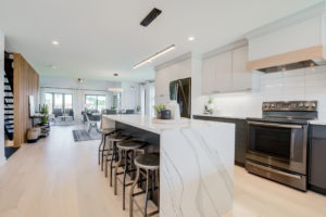Modern kitchen with a large marble island, black barstools, stainless steel appliances, white cabinets, and light wood floors. The space is bright and open, leading to a dining and living area in the background—an inviting setting that reflects the stylish efficiency found in many net zero homes.
