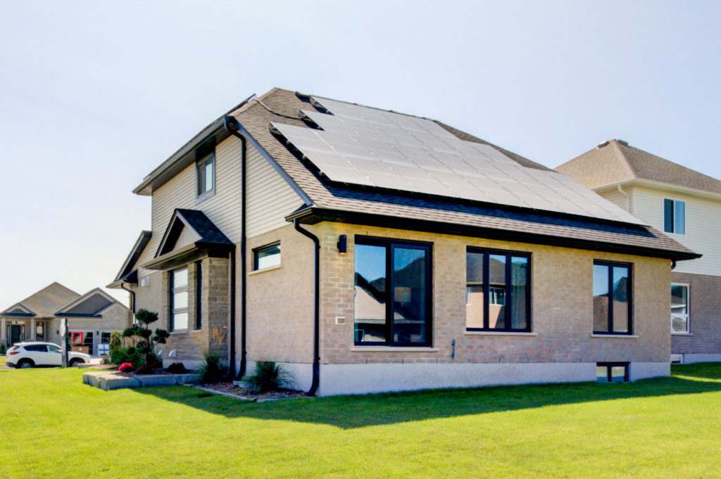 A modern house with beige brick and siding has solar panels installed on the roof and sits on a green lawn in a suburban neighborhood under a clear sky, reflecting the growing trend of net zero homes within net zero communities.