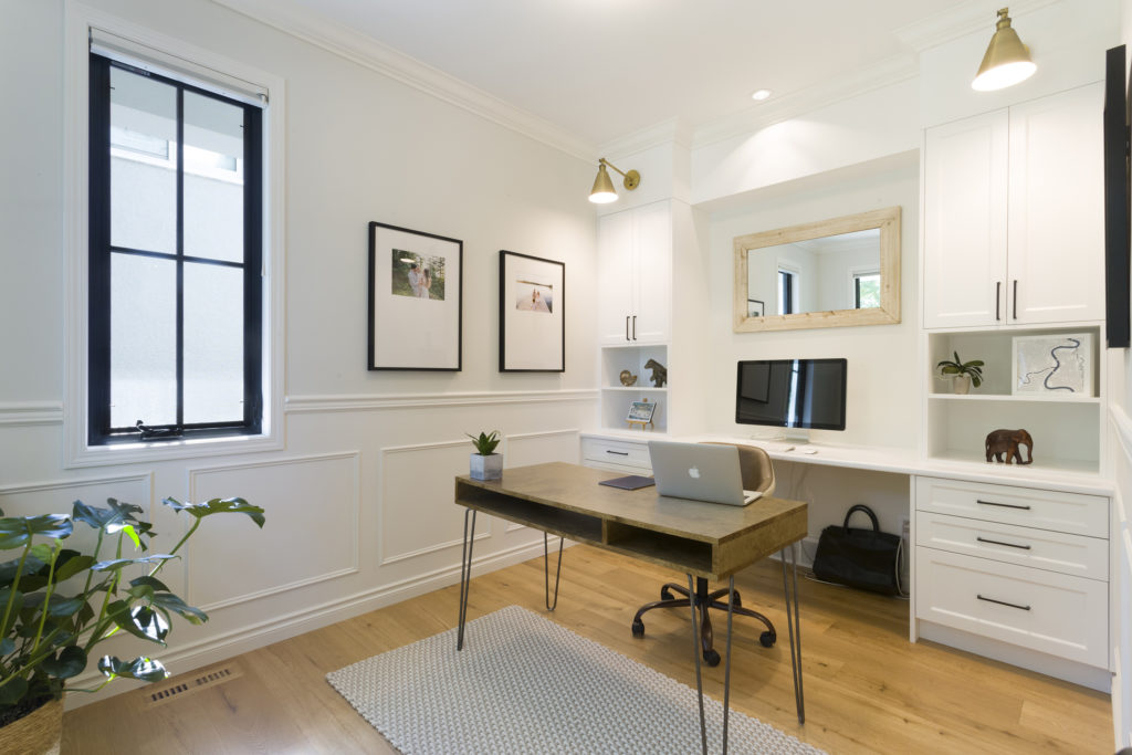 A modern home office designed to meet your home needs features a wooden desk, laptop, desktop monitor, wall art, white cabinets, a large mirror, an indoor plant, and light wood flooring.