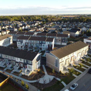 Aerial view of a suburban neighborhood with rows of modern townhouses, small yards, sidewalks, and green spaces—part of one of the region’s emerging net zero communities—surrounded by more homes and trees under a clear sky.