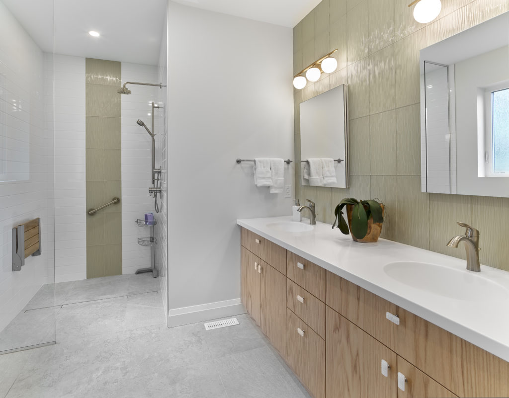 Modern bathroom with double sink vanity, light wood cabinets, a potted plant, wall-mounted mirrors, and a walk-in shower equipped for accessibility at home with a fold-down bench and grab bars. Towel rack with white towels on the wall meets both your practical home needs and modern design preferences.
