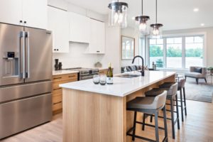 Modern kitchen with a large island, three stools, pendant lights, stainless steel fridge, white cabinets, and large windows letting in natural light—an ideal space for net zero homes striving for sustainability within net zero communities.