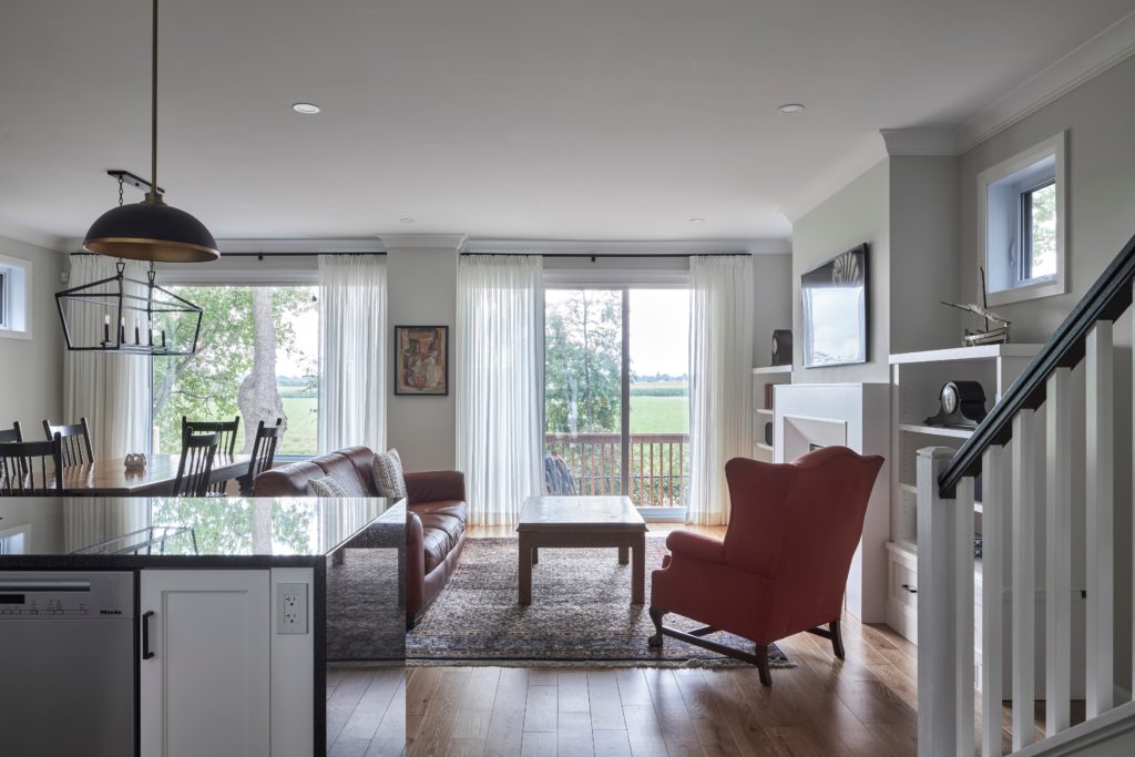 Bright living room with a red armchair, sofa, and coffee table on a rug, dining area to the left, large windows with white curtains, and a view of trees and fields outside. Stairs and kitchen are visible in the foreground—an inviting space designed for comfort in one of our thoughtfully planned net zero homes.