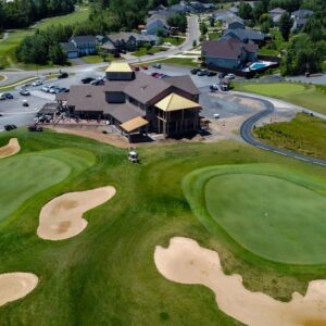 Aerial view of a golf course with sand bunkers, two putting greens, and a large clubhouse surrounded by parked cars and residential houses—including eco-friendly net zero homes—in the background.