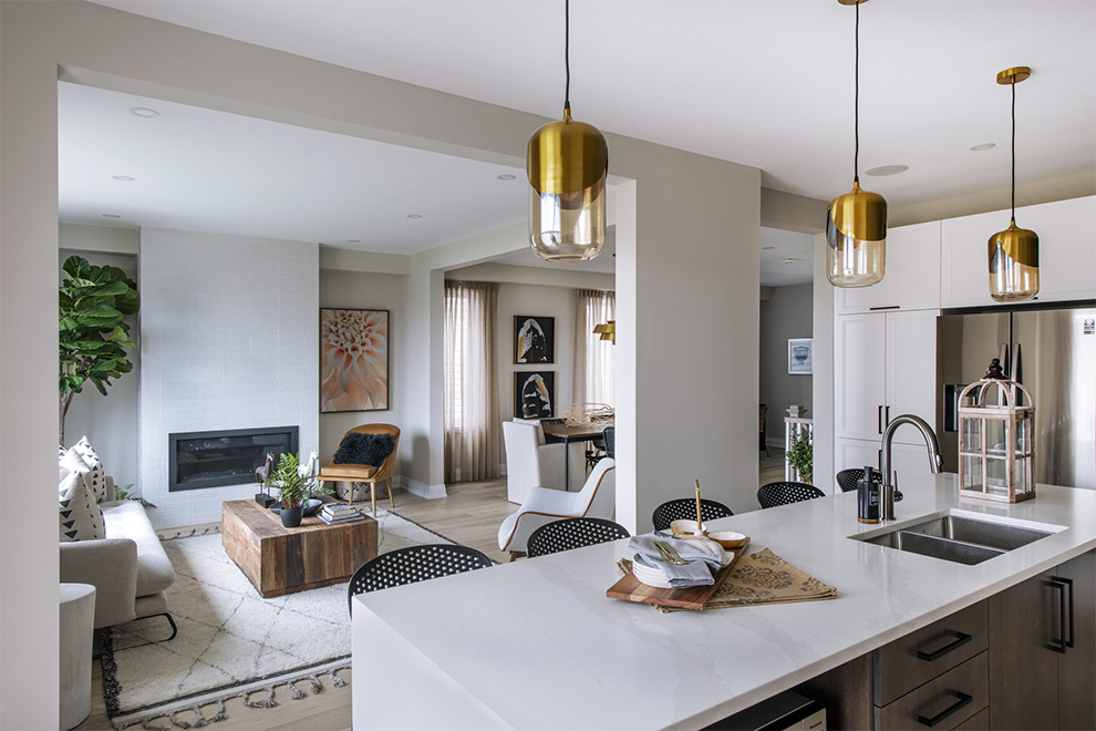 Modern open-concept kitchen and living room with white countertops, gold pendant lights, a gray sofa, fireplace, wooden coffee table, and large windows letting in natural light—perfect for those seeking stylish comfort in today’s innovative net zero homes.