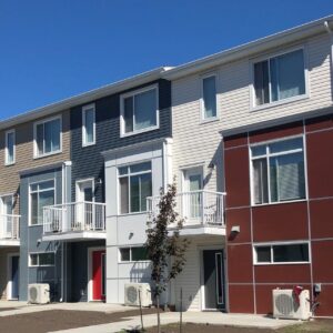A row of modern, three-story townhouses with colorful facades and small balconies, set against a clear blue sky. Designed as net zero homes, these residences exemplify sustainable living. A young tree is planted in front of the buildings, enhancing the eco-friendly atmosphere of this forward-thinking community.