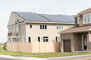 A tan two-story house with solar panels on the roof exemplifies the promise of net zero homes, standing next to a brown house with a garage—both surrounded by wooden fences and a sidewalk, reflecting the growing trend of net zero communities.