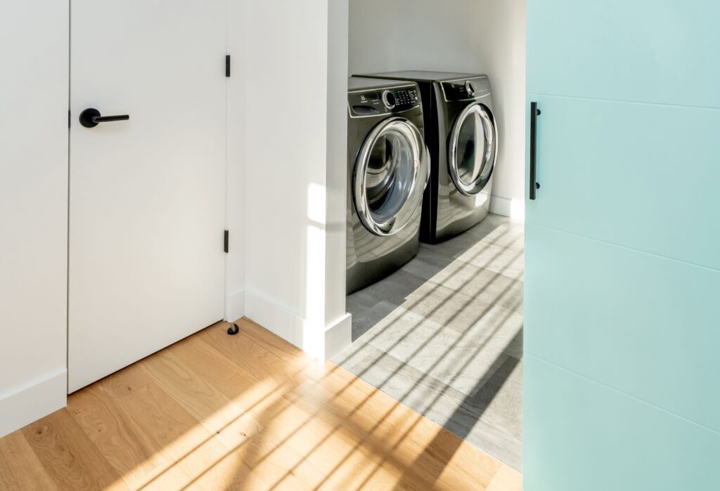 A modern laundry room with a front-loading washer and dryer, white walls, light wood flooring, and sunlight streaming through window blinds—perfect for your Spring Maintenance Inspection to help protect your home as the seasons change, with gentle shadows cast across the floor.