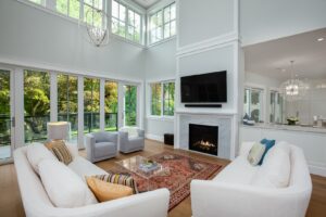 Modern living room with high ceilings, large windows, two white sofas, two armchairs, a red patterned rug, a marble fireplace with a TV above, and a view into the kitchen—an inviting space that reflects Housing Excellence worthy of CHBA National Awards recognition.