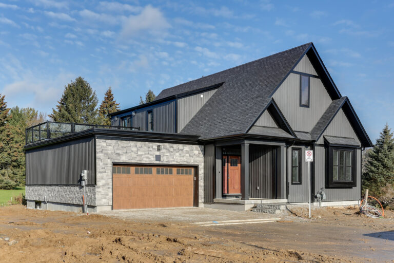 Modern two-story house with dark siding, stone accents, and a wooden garage door. The yard is unfinished dirt. Pine trees and a blue sky are visible in the background—an inviting scene reminiscent of homes recognized by the CHBA National Awards for Housing Excellence.