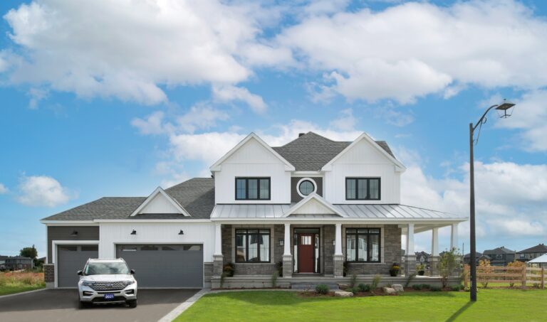 A modern two-story house, recognized for Housing Excellence, features a gray roof, white siding, a red front door, a wraparound porch, and a spacious three-car garage. A white SUV is parked in the driveway, and the yard is neatly landscaped under a blue sky—an inviting example that reflects standards celebrated by the CHBA National Awards for Housing Excellence.