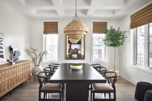 Modern dining room with a dark table, eight black chairs, a wood sideboard, large mirror, potted tree, two windows with brown shades, and a layered rattan chandelier. Decor includes vases and a bowl on the table—a sophisticated space that reflects the style celebrated by the CHBA National Awards for Housing Excellence.