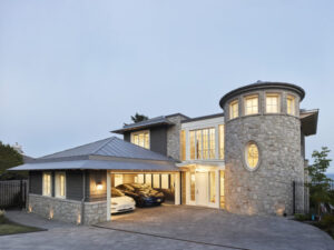 Modern two-story house with a stone turret, large windows, and three cars parked in an open garage. The house is lit warmly from within, and the driveway is paved with gray bricks—a striking example of Housing Excellence recognized by the CHBA National Awards for Housing Excellence.