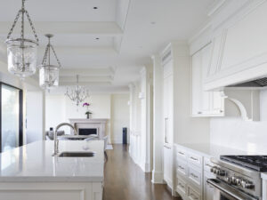 Modern white kitchen with marble countertops, stainless steel appliances, pendant lights, and hardwood floors. Open layout leads to a living area with a fireplace and a vase with purple flowers on the mantel—a stunning example of Housing Excellence worthy of CHBA National Awards recognition.