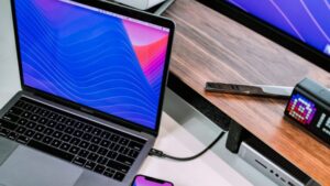 A MacBook with a colorful wavy wallpaper is connected to an energy efficient docking station on a wooden desk, accompanied by a smartphone, monitor, and a digital clock displaying the Instagram logo nearby.
