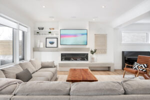 A modern living room with light gray sofas, a wooden coffee table, a wall-mounted TV above a sleek fireplace, shelves with decor, and large windows letting in natural light—an inviting space that exemplifies Housing Excellence as celebrated by the CHBA National Awards.