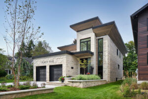 Modern two-story house with stone exterior, large windows, two black garage doors, and FORTY FIVE above the garages—a true example of Housing Excellence. Green lawn, young tree, and potted plants in front. Neighboring houses visible on either side.
