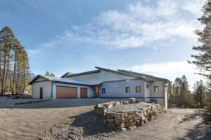 A modern house with gray siding and a three-car garage sits on a sloped gravel driveway, surrounded by pine trees and a blue sky. A low stone retaining wall is in the foreground, reflecting the superior design often recognized by the CHBA National Awards for Housing Excellence.