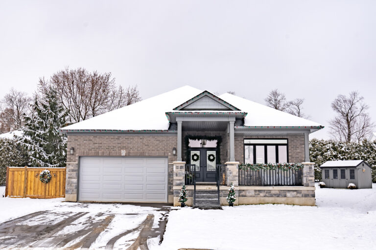 A single-story modern house with a two-car garage and front porch, decorated with holiday wreaths and garland. Snow covers the roof, yard, and trees, creating a winter scene reminiscent of homes featured in the CHBA National Awards for Housing Excellence.