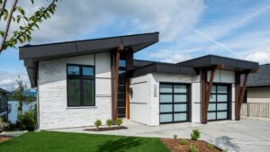 A modern net zero home with white stone walls, large windows, two glass garage doors, and a sloped roof stands on a manicured lawn under a partly cloudy sky. The superior building envelope ensures energy efficiency while blending contemporary aesthetics with sustainable living.