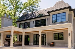 Modern two-story house with large windows, a covered patio area, and outdoor seating. Trees surround the home, and the exterior features light-colored walls and a dark roof. Designed with home energy efficiency in mind, this residence offers contemporary comfort while embodying standards recognized by the National Awards for Housing Excellence.
