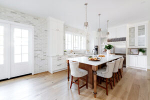 Bright, modern kitchen with white cabinets, marble island with wooden base, four cushioned chairs, pendant lights, stainless steel appliances, and light wood floors. Natural light streams through large windows and glass doors. Designed with home energy efficiency in mind, this space combines style and sustainability for an award-worthy living experience.