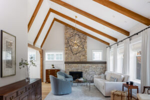 Bright living room with vaulted ceiling, exposed wooden beams, stone fireplace, white sofa, blue armchairs, large windows with curtains, and modern geometric chandelier—designed to maximize home energy efficiency while achieving a style worthy of National Awards for Housing Excellence.