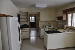 A simple kitchen with cream cabinets, a refrigerator, stove, dishwasher, and a sink. There’s a window with shutters, shelves, and a doorway leading to another room. The countertops have a few items placed on them—designed with home energy efficiency in mind for modern living.