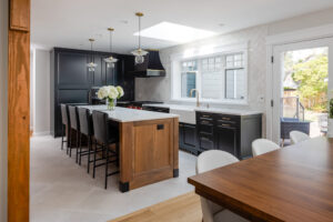 Modern kitchen with black cabinets, a large wooden island with white countertop, bar stools, pendant lights, a dining table, and a window letting in natural light. Designed with home energy efficiency in mind, this inviting space opens to a patio where outdoor greenery is visible—a quality often recognized by the National Awards for Housing Excellence.