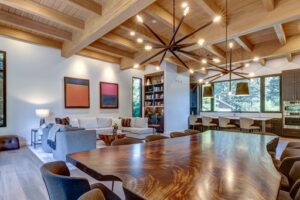 A modern open-concept living and dining room with wood ceilings, a large wood table, stylish light fixtures, white sofas, wall art, bookshelves, and large windows letting in natural light—designed with home energy efficiency in mind and inspired by the standards of the National Awards for Housing Excellence.