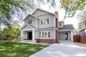 Two-story gray house with white trim, red brick accents, large front windows, a covered entry, and a spacious driveway. Green lawn and trees surround the home, creating a welcoming suburban atmosphere that highlights home energy efficiency throughout its design.