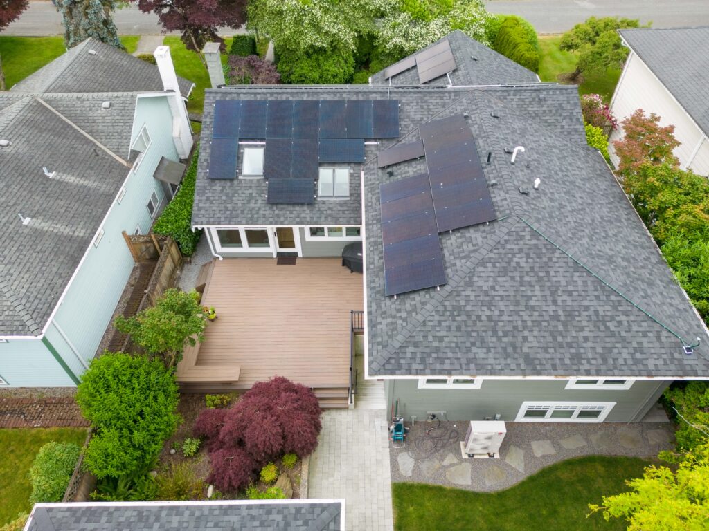 Aerial view of a house with solar panels on the roof, showcasing home energy efficiency, a large wooden deck, surrounding green trees and shrubs, and neighboring houses visible on both sides.