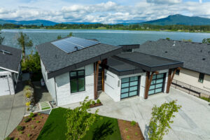 Modern house with solar panels on the roof, large windows, and a glass garage door, designed with home energy efficiency in mind, situated beside a lake with mountains and greenery in the background.