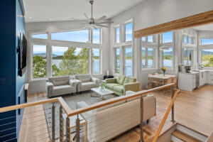 Modern living room with large windows, natural light, lake views, light wood floors, neutral furniture, and open kitchen—designed with home energy efficiency in mind. Stairs with metal railing lead down from the entrance. Dining table visible near the windows.
