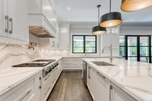 Modern white kitchen with marble countertops, stainless steel appliances, dark wood floor, large island with sink, and two black pendant lights hanging above. Large windows provide natural light while enhancing home energy efficiency.