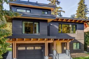 Modern two-story house with dark wood siding, large windows, and a single-car garage. The front entrance has a covered porch supported by wooden beams, and the home is surrounded by tall trees. Designed with home energy efficiency in mind, this residence combines contemporary style with eco-friendly living—a combination that's been recognized at the National Awards for Housing Excellence.