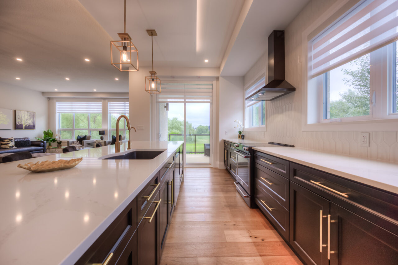 Modern kitchen with black cabinets, white countertops, pendant lights, and large windows offering a view of greenery outside. Open layout connects kitchen to living area with natural light filling the space—an elegant example often found in thoughtfully designed net zero homes.