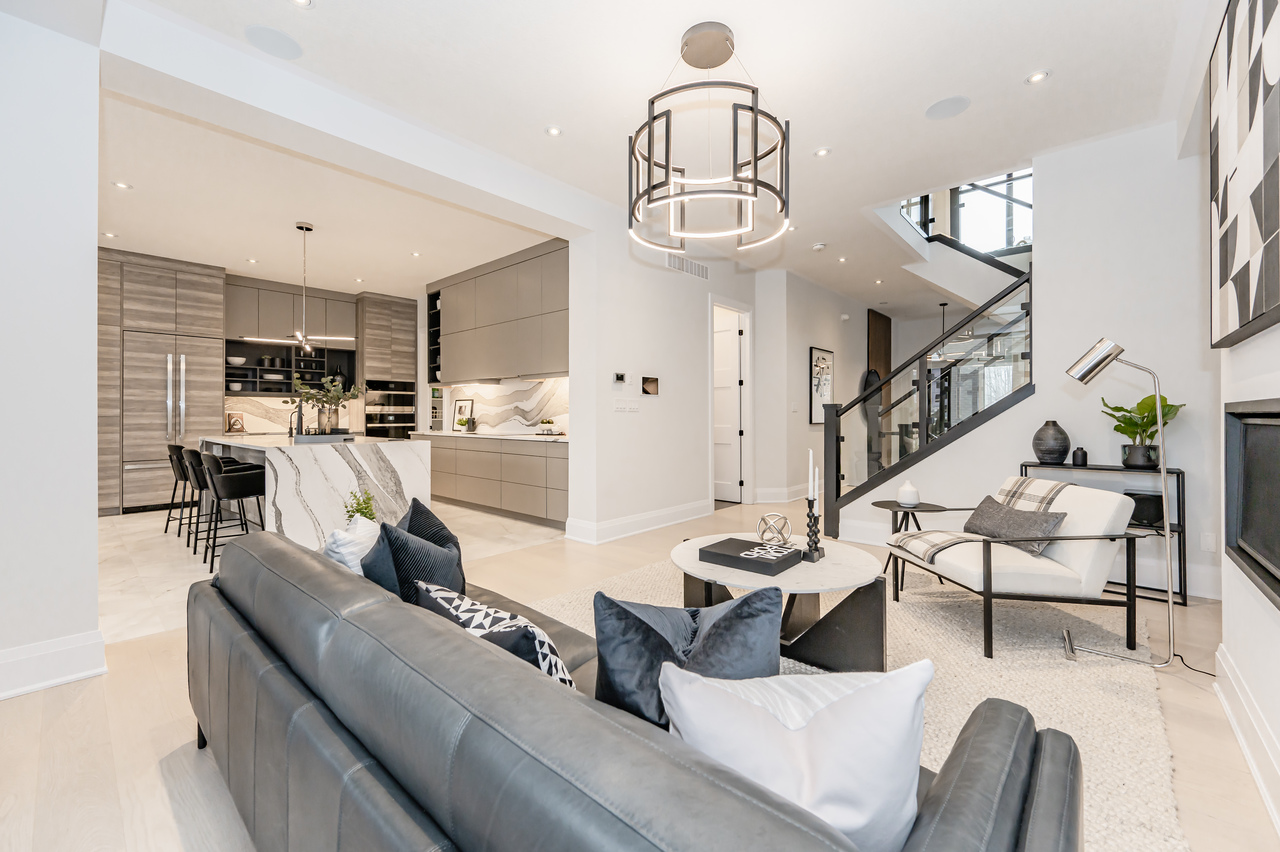 Modern open-concept living space with a gray sofa, stylish lighting fixtures, sleek kitchen with barstools, and stairs with glass railing. Light wood floors and neutral decor create a bright, airy atmosphere—perfectly reflecting the sustainable elegance found in today’s production net zero homes.