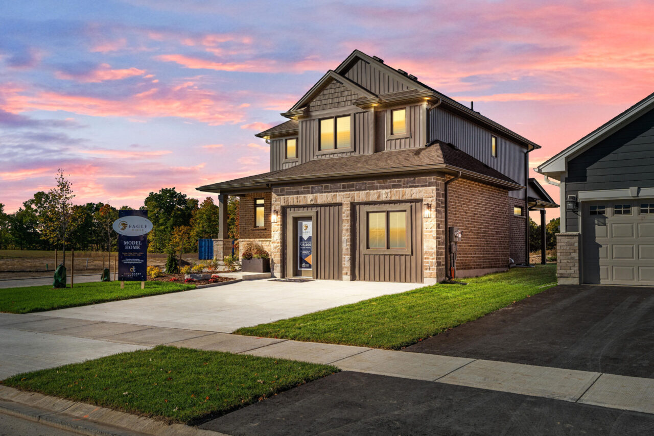 A modern two-story house with brick and siding exterior is shown at sunset. The driveway and lawn are neatly maintained, and a sidewalk runs in front. A sign near the house indicates it is a model home, showcasing the latest advancements in production net zero homes.