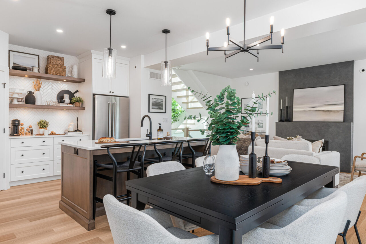 Modern open-concept kitchen and dining area with white cabinets, wood accents, black dining table, and a vase with greenery. Pendant lights hang over the island; a cozy living room is visible in the background—all thoughtfully designed within a net zero home that seamlessly blends style and sustainability.