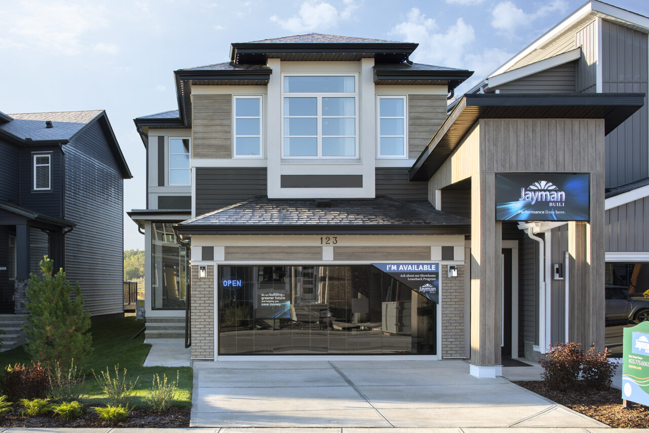 Modern two-story house with large windows, neutral siding, and a garage. A sign on the garage door says IM AVAILABLE, and another sign above the entrance reads Jayman BUILT. Showcasing the latest in production net zero homes, this residence is surrounded by green landscaping along the driveway.