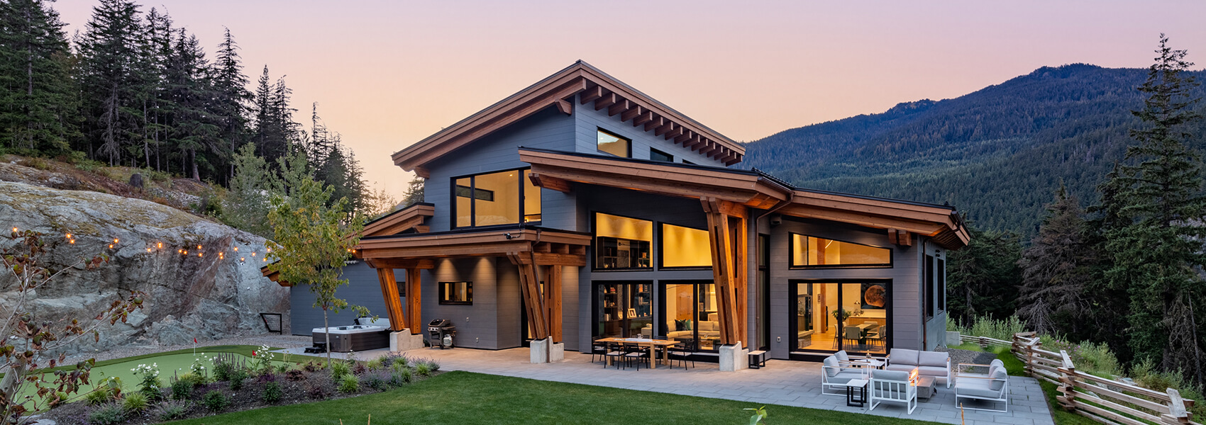 Modern mountain house crafted by Canadian Home Builders' Association, with large wooden beams, tall windows glowing with warm light, surrounded by pine trees, rocky landscape, and patio seating at sunset with stunning mountain views.