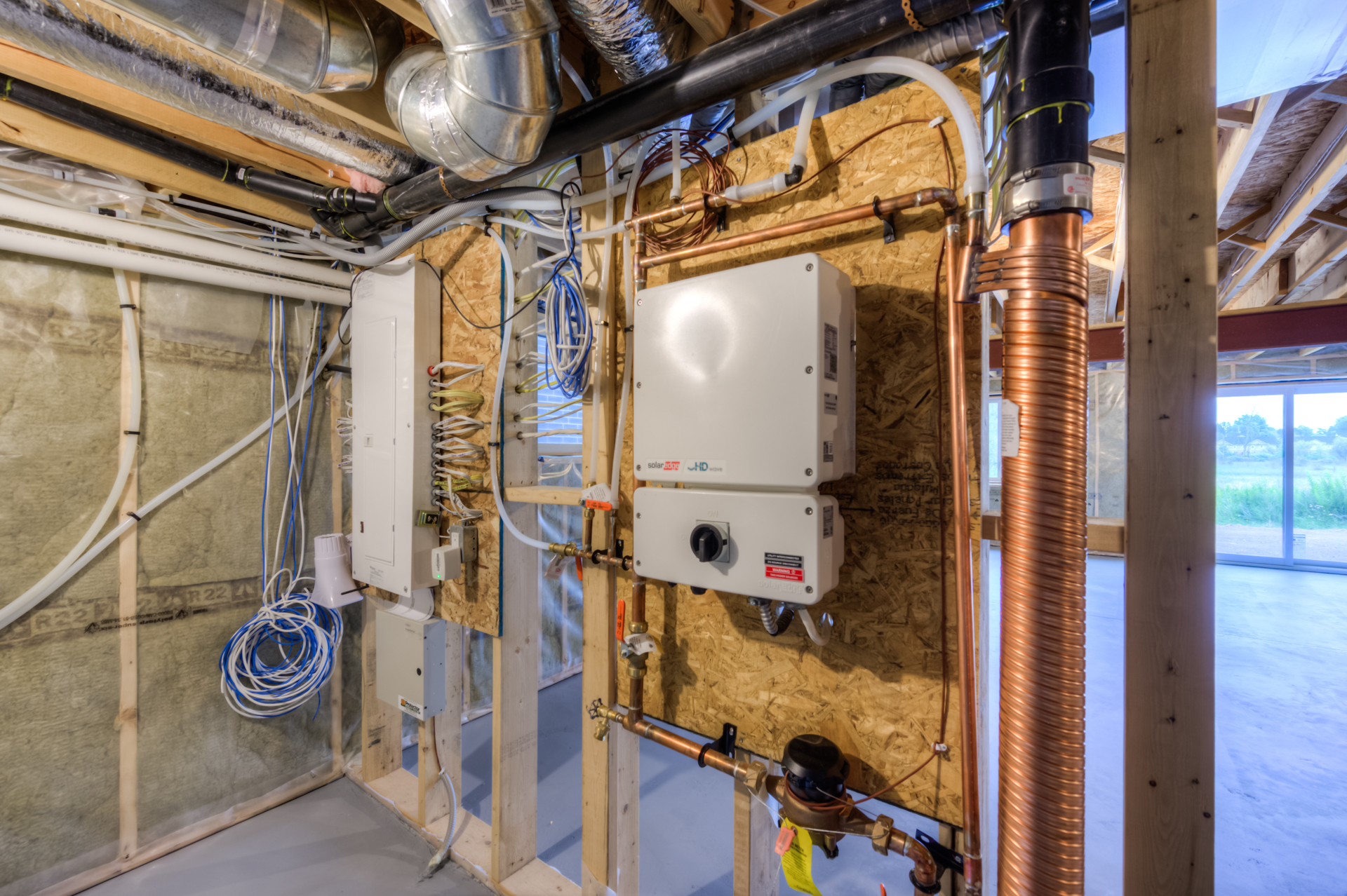 A partially finished basement with exposed framing, insulation, and HVAC ducts highlights the essential infrastructure found in many net zero homes. Mounted on the wall are electrical panels, wiring, and plumbing components—including copper pipes and water heaters—showcasing the attention to detail necessary for production net zero homes.