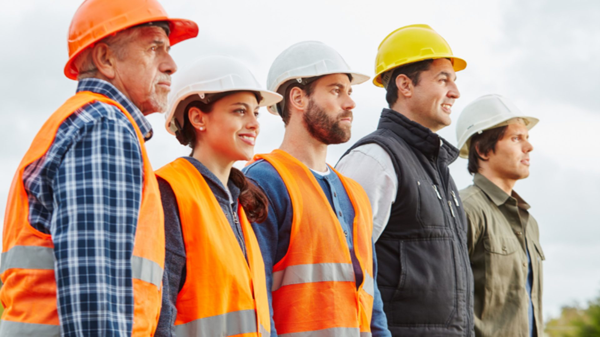 Five construction workers wearing safety vests and helmets stand in a row outdoors, looking ahead with determined expressions. Reflecting the diversity of those pursuing a career in construction, the group includes both men and women.