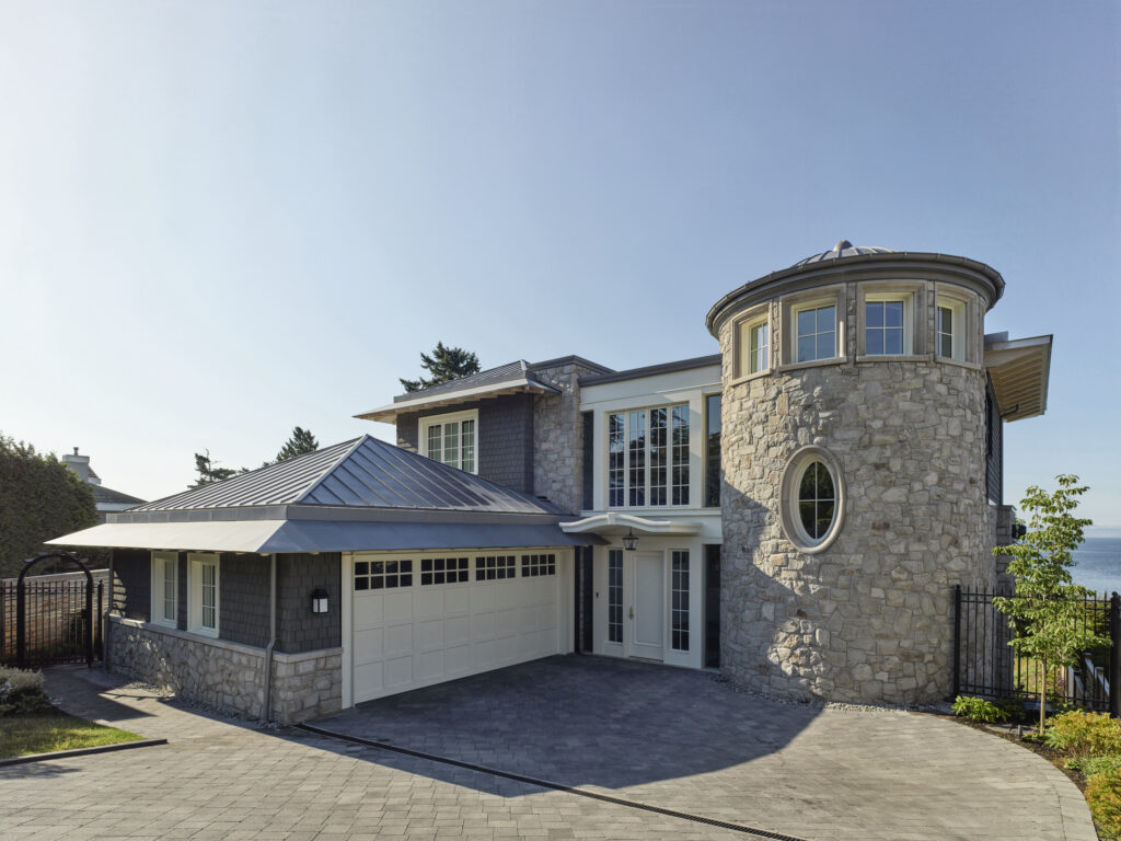 Modern house with stone and shingle exterior, round turret feature, large windows, attached two-car garage, and paved driveway. Designed with the principles of energy efficient homes in mind, this residence blends style and sustainability. Trees and water are visible in the background under a clear blue sky.