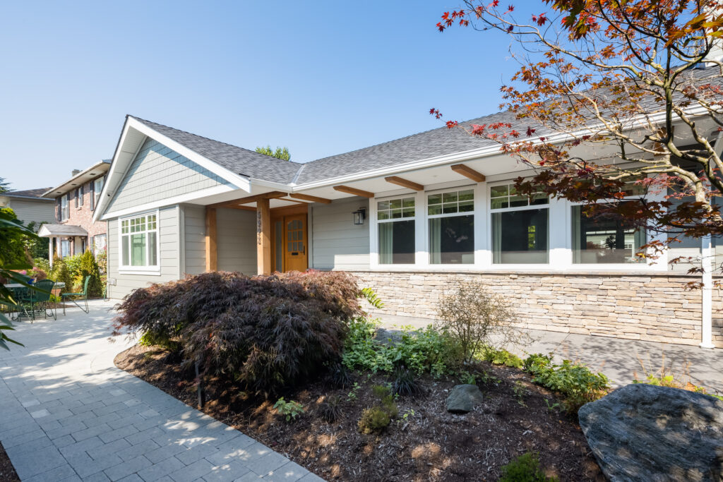 Single-story gray house with a stone accent exterior, large windows, and wooden entrance. This energy efficient home features well-kept landscaping with shrubs and a tree, set under a clear, blue sky.
