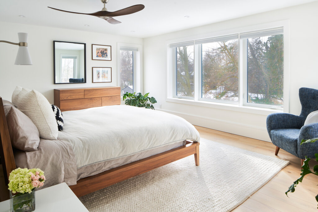 Bright bedroom with large windows, a wooden bed and dresser, white bedding, a blue armchair, green plants, framed art on the wall, and light wood flooring. This inviting space combines style and sustainability—perfect for those who appreciate energy efficient homes. A bouquet of flowers sits on a small white table.