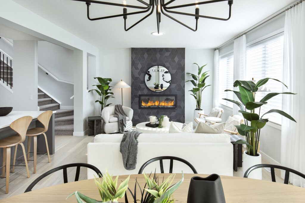 Modern living room with a white sofa, fireplace, round mirror, large windows with sheer curtains, indoor plants, and a dining table with chairs in the foreground. Neutral decor and natural light create a cozy atmosphere—perfect for those seeking energy efficient homes that combine style and sustainability.